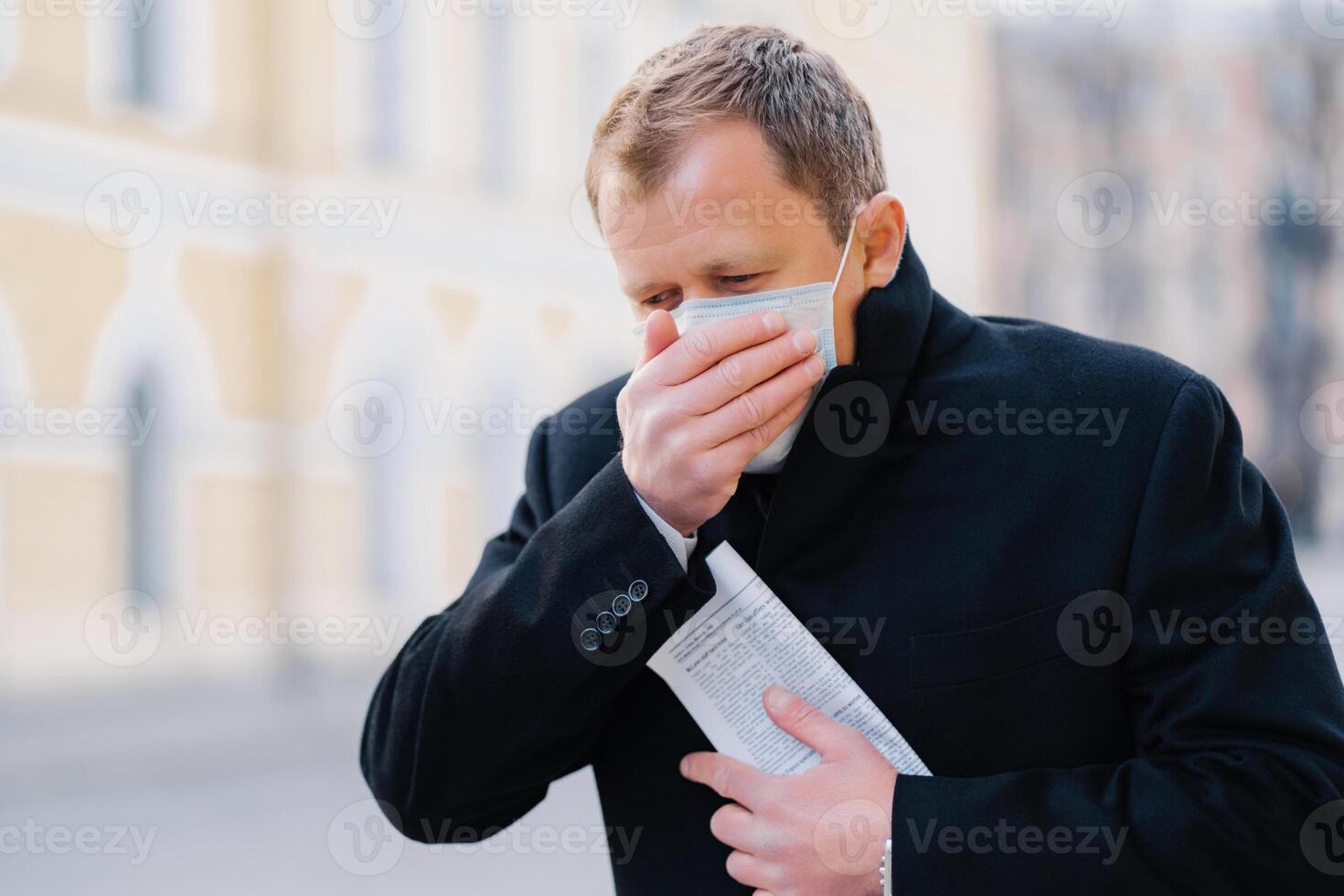 malade homme dans noir manteau couvrant bouche avec masque, en plein air, montrant panneaux de maladie, dans un Urbain paramètre, mise en évidence santé soucis photo