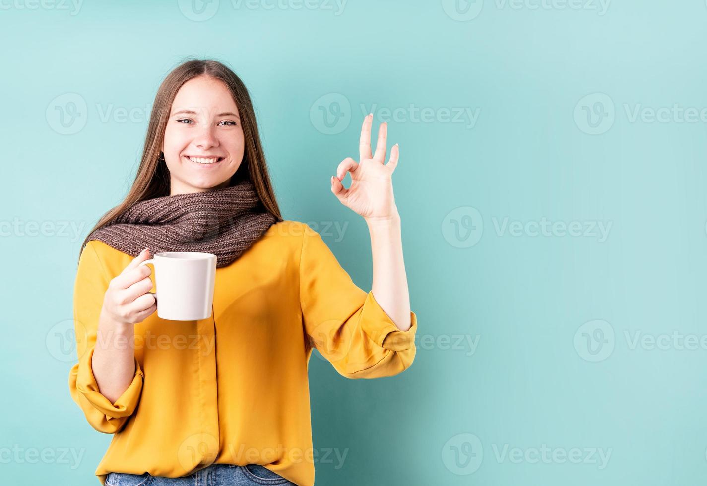 Jeune femme de race blanche portant un foulard buvant du café ou du thé montrant un signe ok sur fond bleu photo