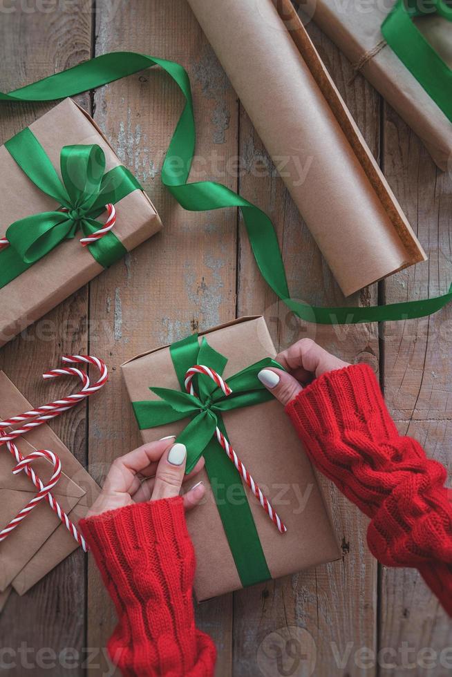 mains de femme tenant un cadeau de noël enveloppé de papier kraft, ruban vert et canne en bonbon vue de dessus sur table en bois photo