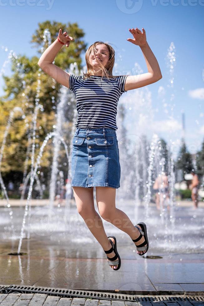 Jeune femme caucasienne séduisante et heureuse sautant et riant près de la fontaine dans le parc urbain photo