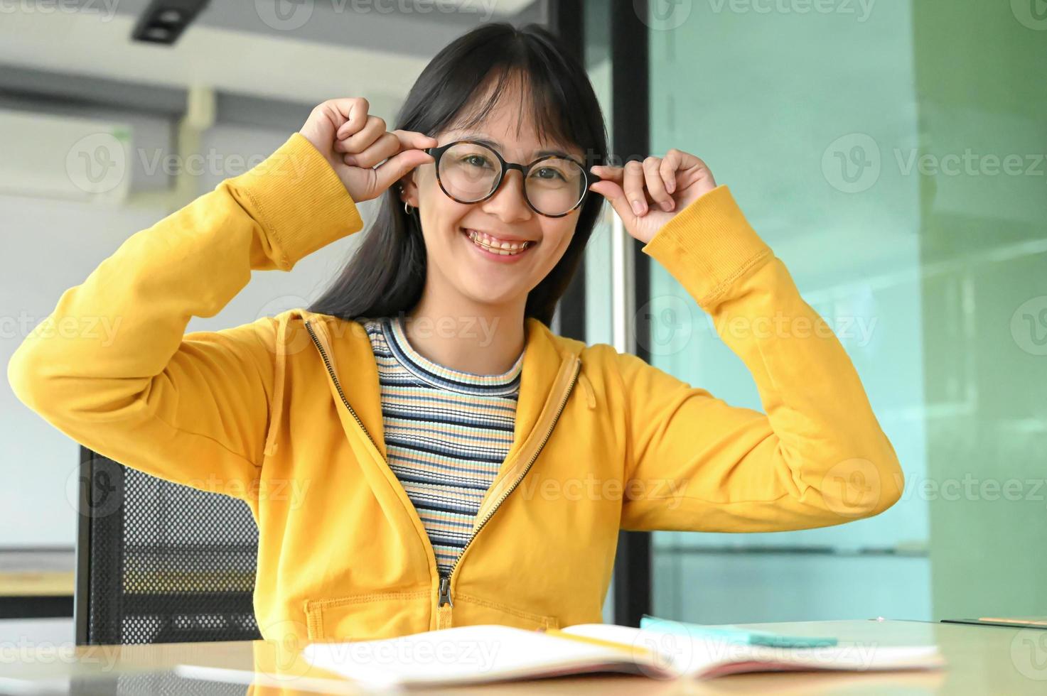 étudiante asiatique avec des lunettes et sourit pour la caméra. elle lit des livres de préparation aux examens. photo