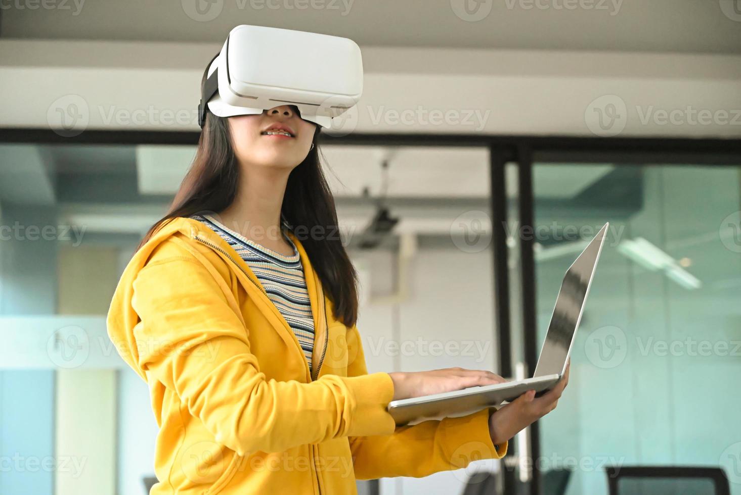 fille asiatique portant une chemise jaune, utilisant un casque vr. elle tient un ordinateur portable et tape un clavier. photo
