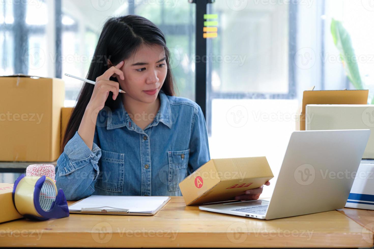 le personnel de livraison des colis recherche le numéro de produit à l'aide d'un ordinateur portable. elle tient un stylo et une boîte à colis à la main. photo