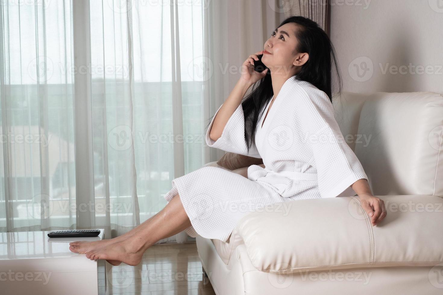 jeune femme asiatique séduisante dans un peignoir assis sur le canapé qui parle au téléphone dans la chambre photo