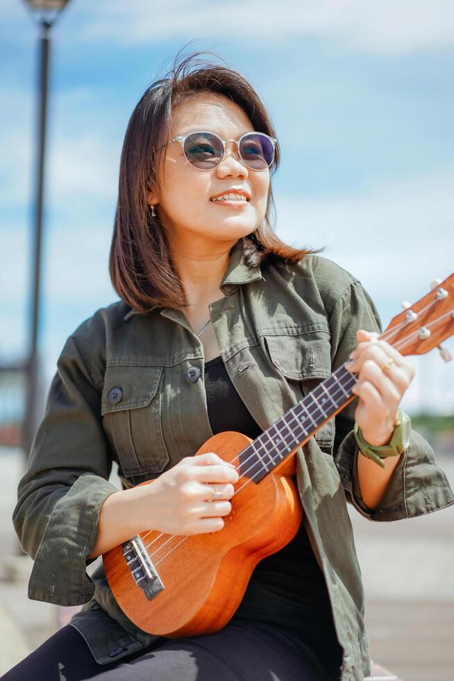 en jouant ukulélé de Jeune magnifique asiatique femme portant veste et noir jeans posant en plein air photo