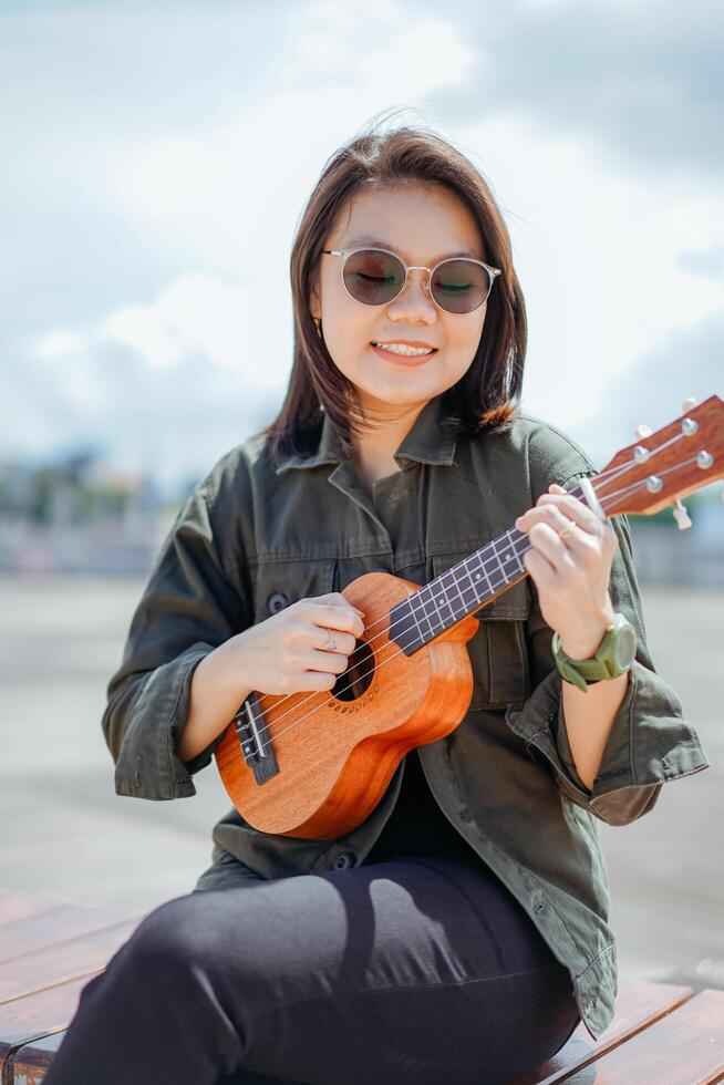 en jouant ukulélé de Jeune magnifique asiatique femme portant veste et noir jeans posant en plein air photo