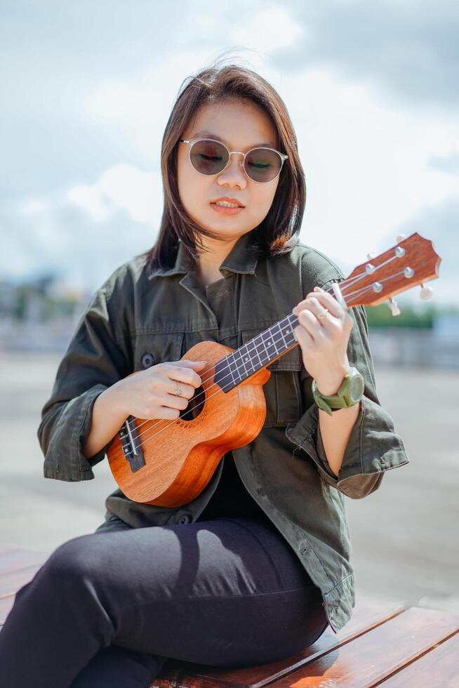 en jouant ukulélé de Jeune magnifique asiatique femme portant veste et noir jeans posant en plein air photo