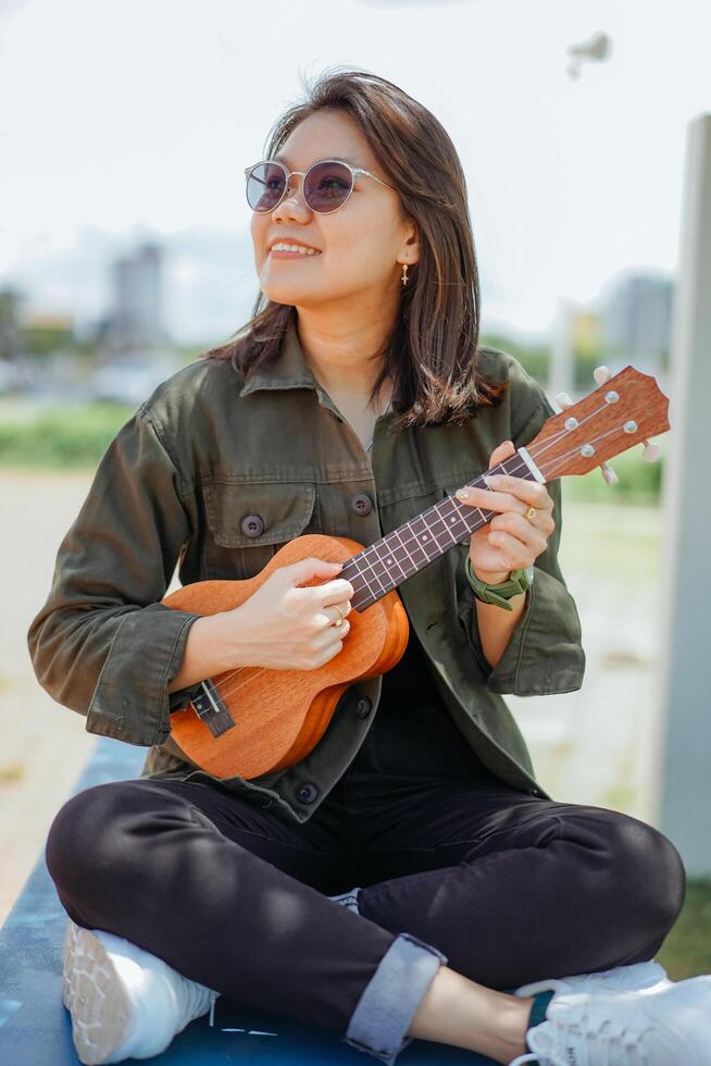 en jouant ukulélé de Jeune magnifique asiatique femme portant veste et noir jeans posant en plein air photo