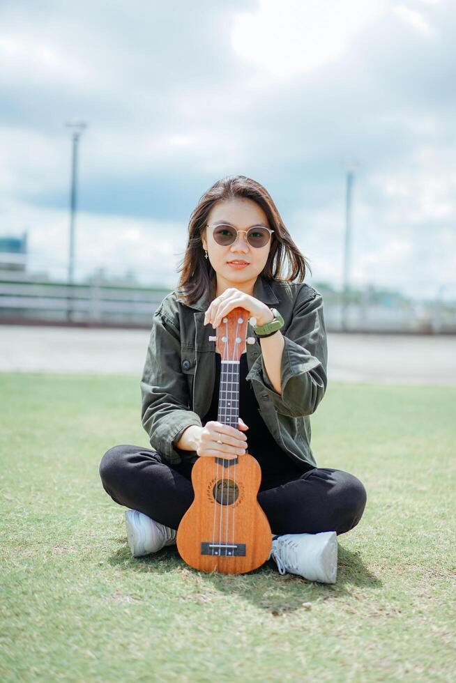 en jouant ukulélé de Jeune magnifique asiatique femme portant veste et noir jeans posant en plein air photo