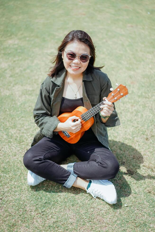 en jouant ukulélé de Jeune magnifique asiatique femme portant veste et noir jeans posant en plein air photo