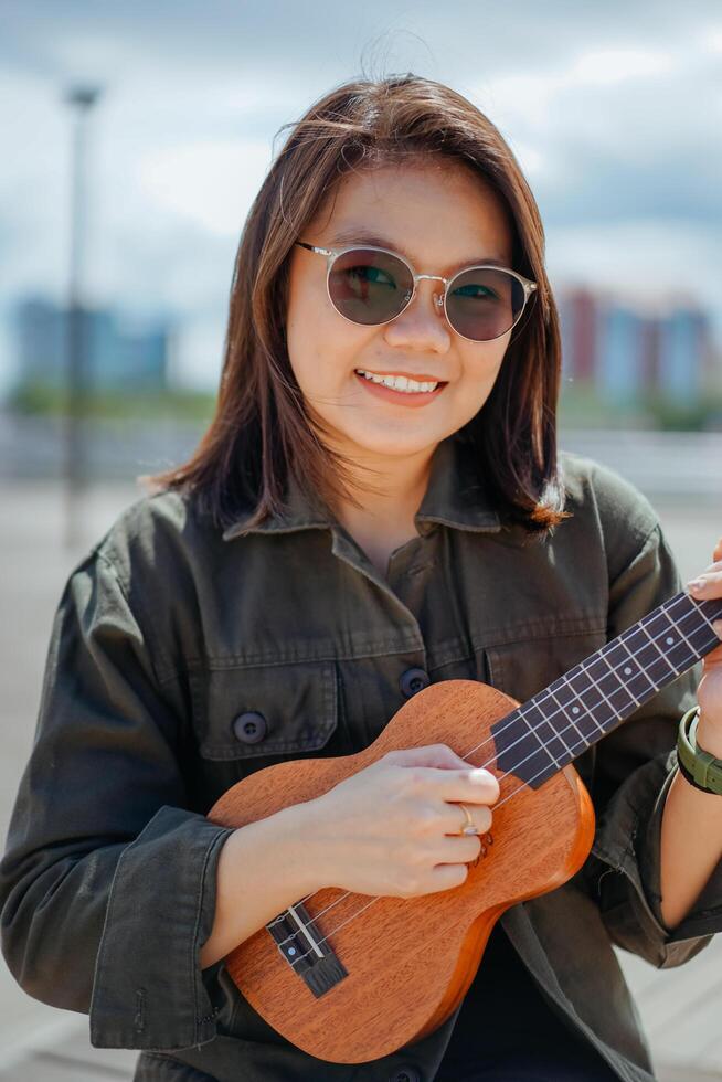 en jouant ukulélé de Jeune magnifique asiatique femme portant veste et noir jeans posant en plein air photo