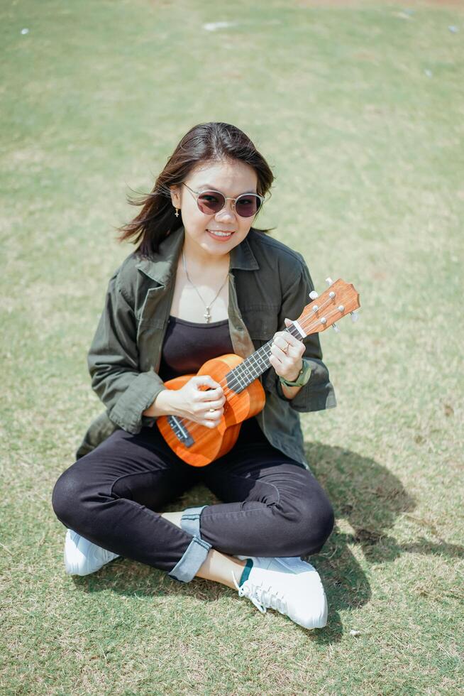 en jouant ukulélé de Jeune magnifique asiatique femme portant veste et noir jeans posant en plein air photo