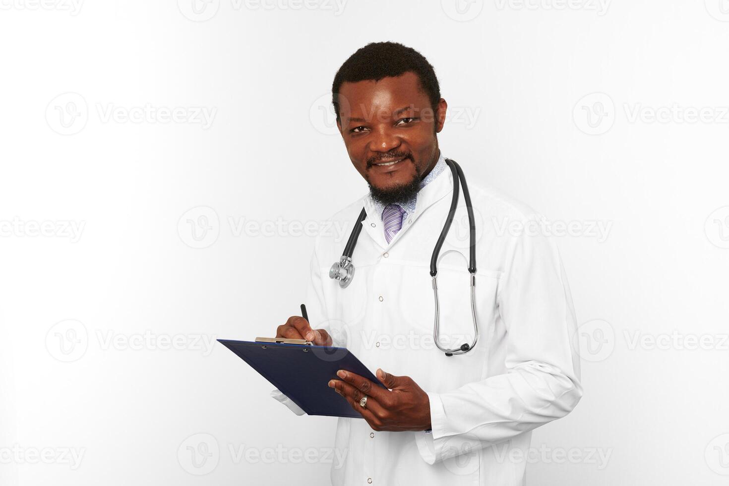 homme médecin barbu noir souriant en robe blanche avec stéthoscope remplissant les dossiers médicaux sur le presse-papiers photo