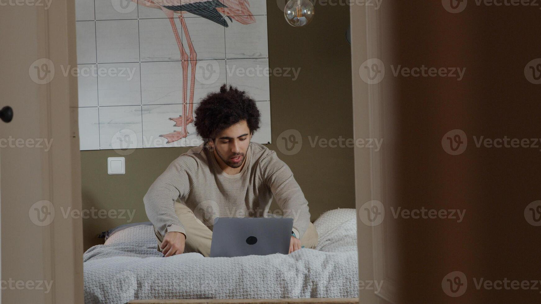 Jeune homme du Moyen-Orient est assis sur le lit, ordinateur portable en face de lui, ayant une conversation en ligne, ajustant les cheveux et parlant photo