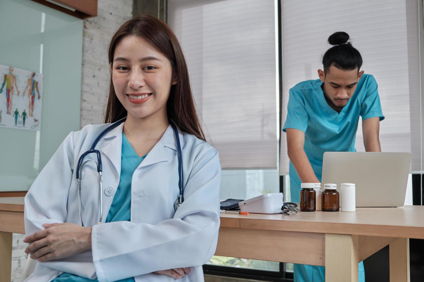 portrait de la belle femme médecin d'origine asiatique en uniforme avec stéthoscope. sourire et regardant la caméra dans une clinique hospitalière, partenaire masculin travaillant derrière elle, deux professionnels. photo