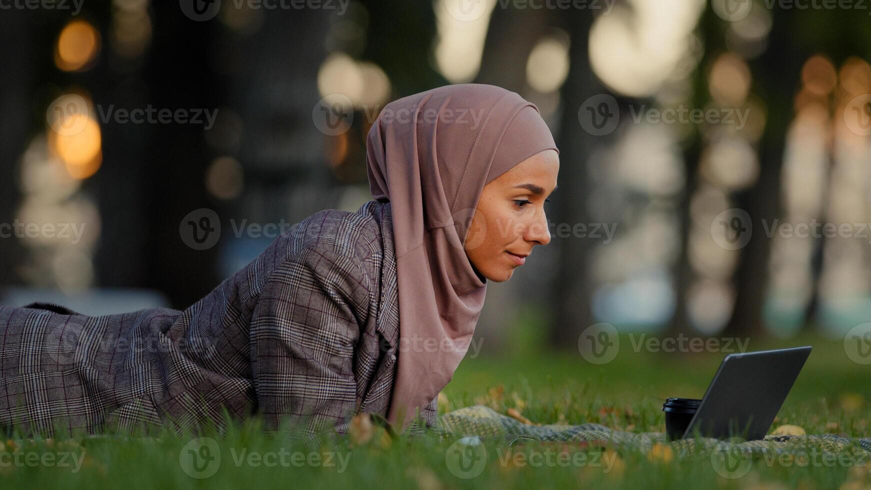 amical islamique affaires femme musulman fille dans hijab mensonge herbe pelouse dans parc en plein air femelle prof en ligne mentor fabrication vidéo appel bavarder conférence parlant webcam en utilisant portable loin la communication photo