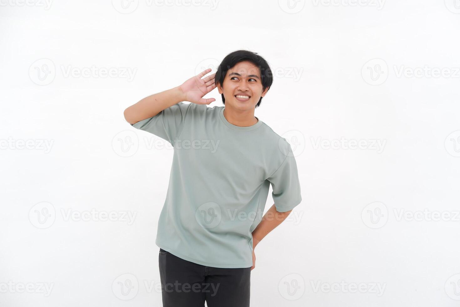 une Jeune asiatique homme, un employé dans une T-shirt, est vu avec enthousiasme écoute avec le sien main à le sien oreille contre une blanc Contexte. photo