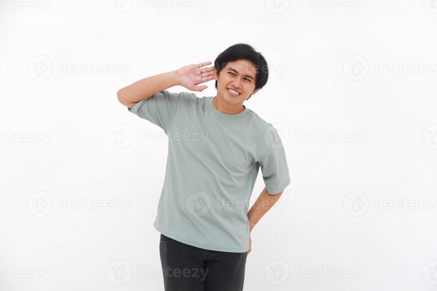 une Jeune asiatique homme, un employé dans une T-shirt, est vu avec enthousiasme écoute avec le sien main à le sien oreille contre une blanc Contexte. photo