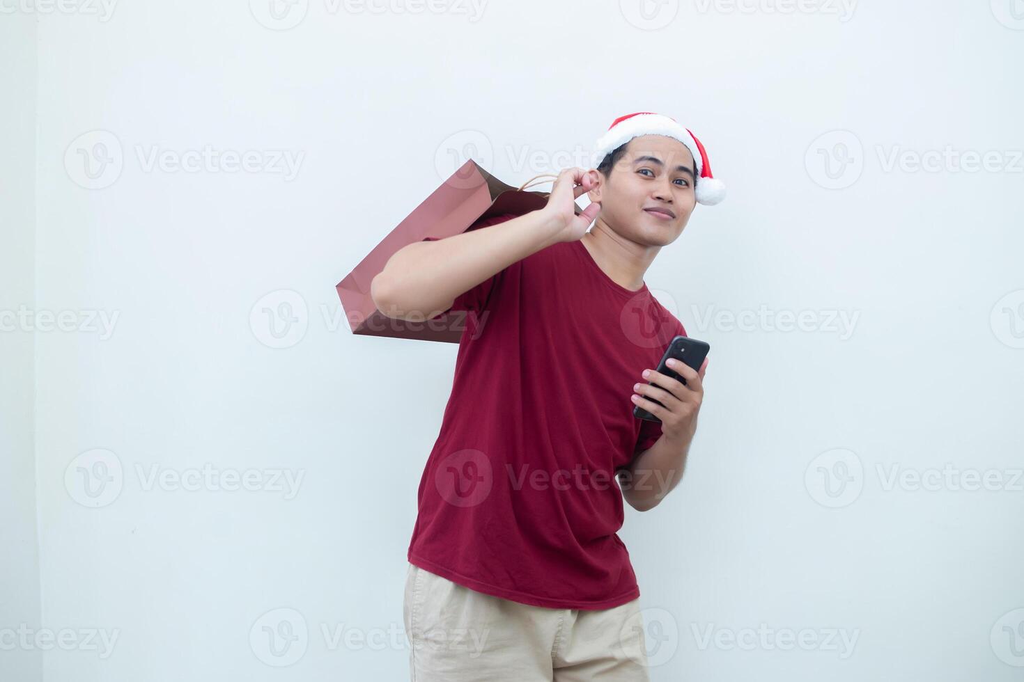 Jeune asiatique homme portant une Père Noël claus chapeau en portant une téléphone intelligent et une achats sac avec expressions de sourire, choc, et surprendre, isolé contre une blanc Contexte pour visuel la communication photo