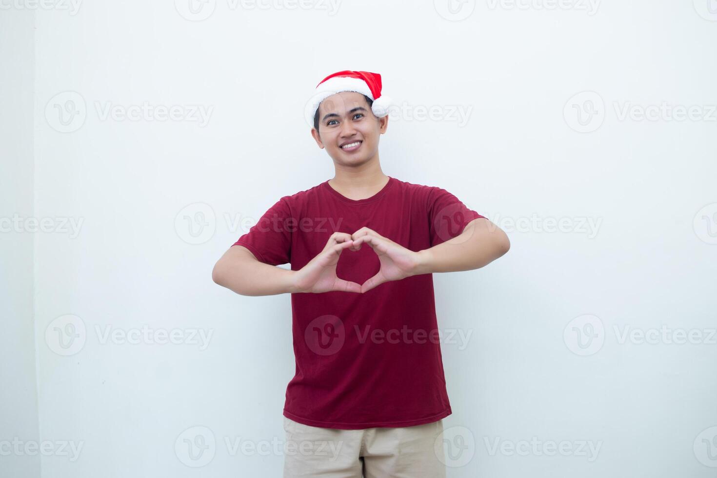 Jeune asiatique homme portant une Père Noël claus chapeau exprimer l'amour par formant une cœur symbole avec le sien mains isolé par une blanc Contexte pour visuel la communication photo