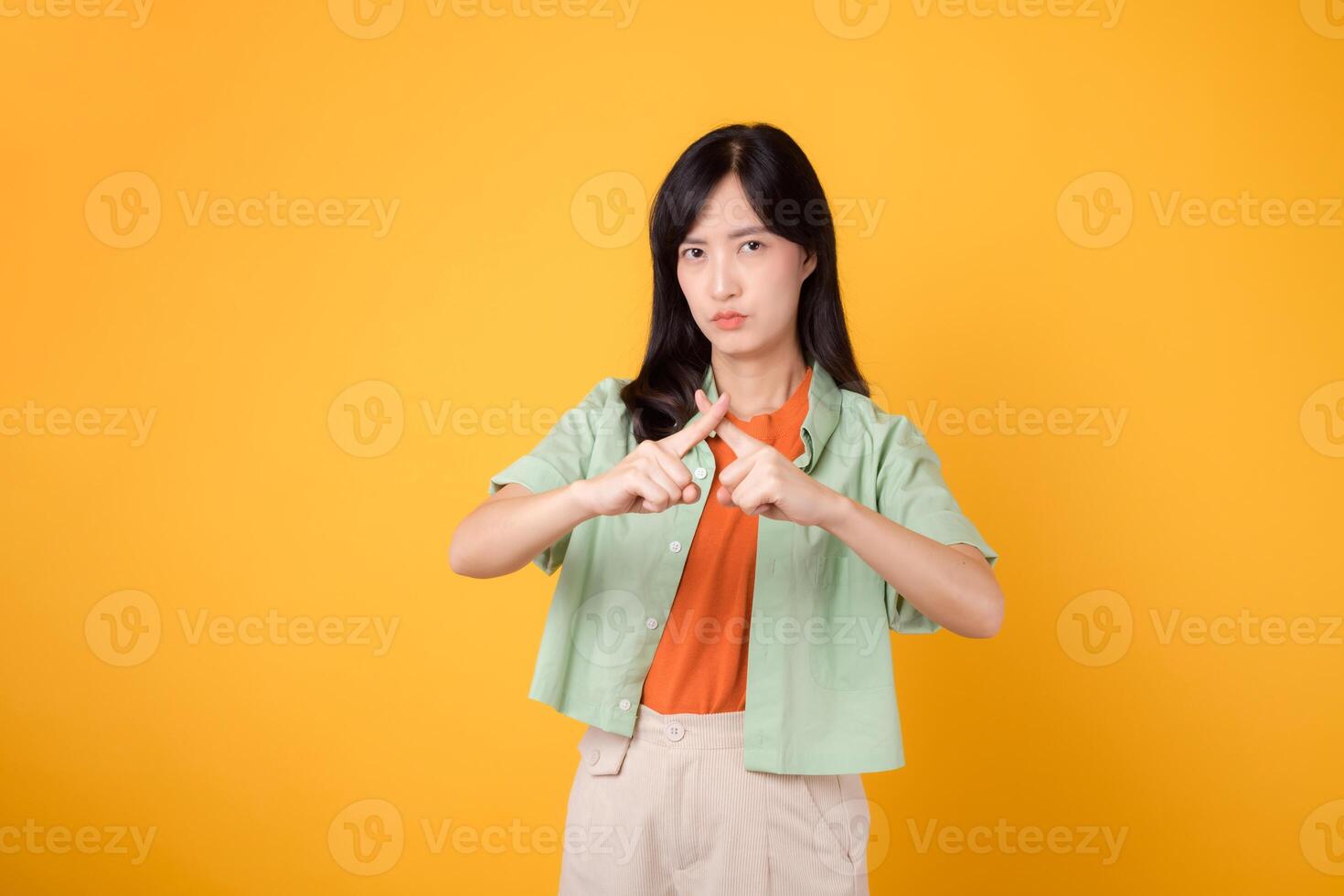 le déni avec une Jeune asiatique femme 30s, élégamment habillé dans Orange chemise et vert sauteur. sa traverser main geste, isolé sur vibrant Jaune arrière-plan, représente concept de refus et désaccord. photo