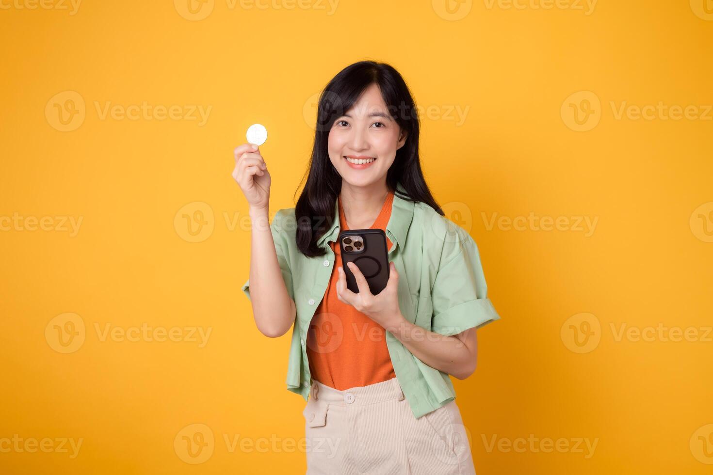 content Jeune asiatique femme dans sa 30s, portant Orange chemise et vert sauteur, vitrines téléphone intelligent écran afficher et crypto devise pièce de monnaie sur Jaune Contexte. futur la finance concept. photo