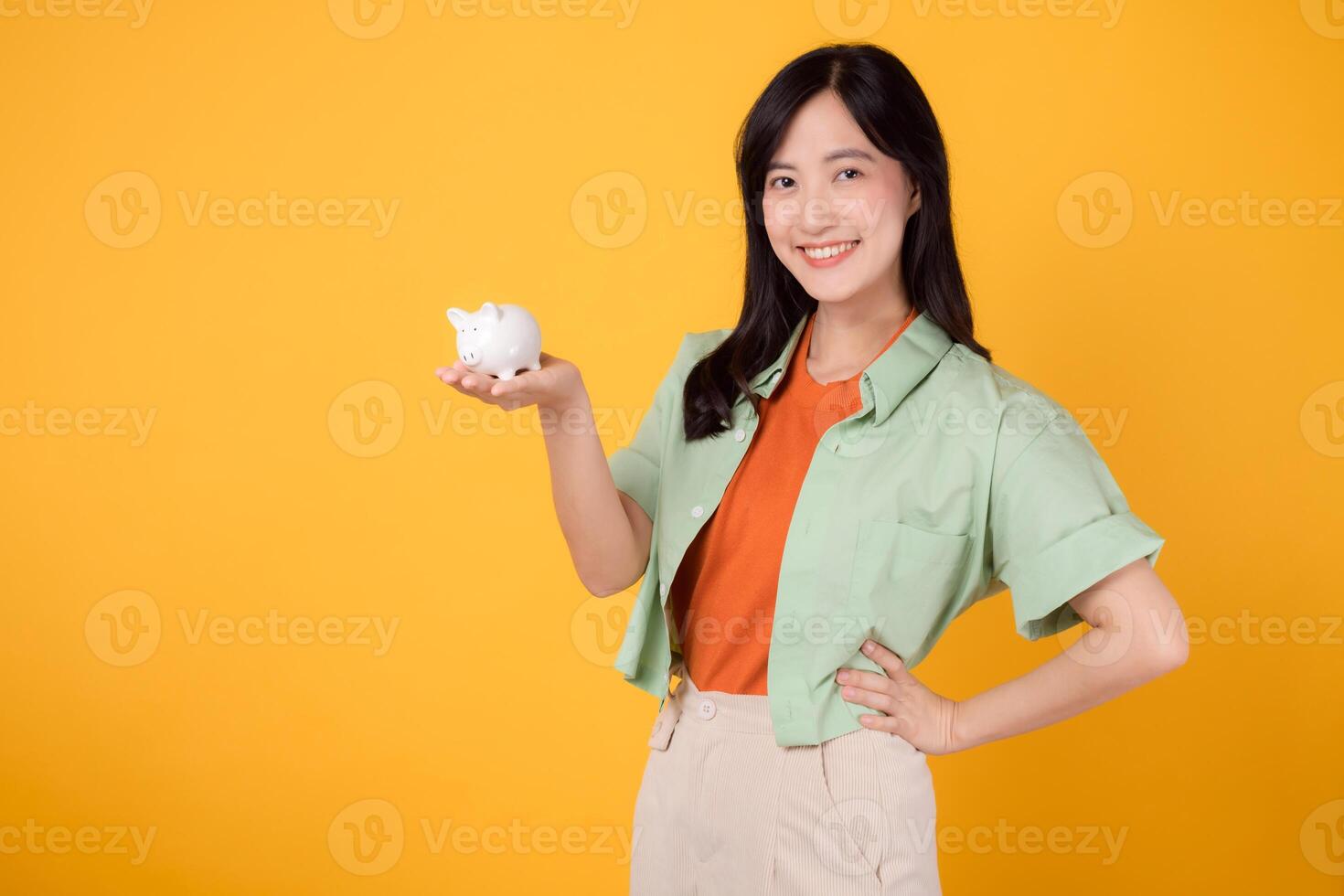 financier argent avec de bonne humeur Jeune asiatique femme dans sa 30s, enfiler Orange chemise et vert sauteur, affichage porcin banque tandis que frappant Akimbo geste sur Jaune Contexte. financier argent concept. photo