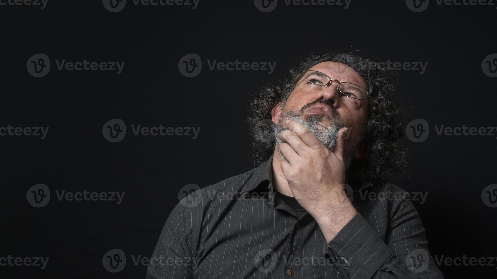 homme avec blanc barbe et noir frisé cheveux avec expression de indécision et doute, à la recherche en haut, portant noir chemise contre noir Contexte photo