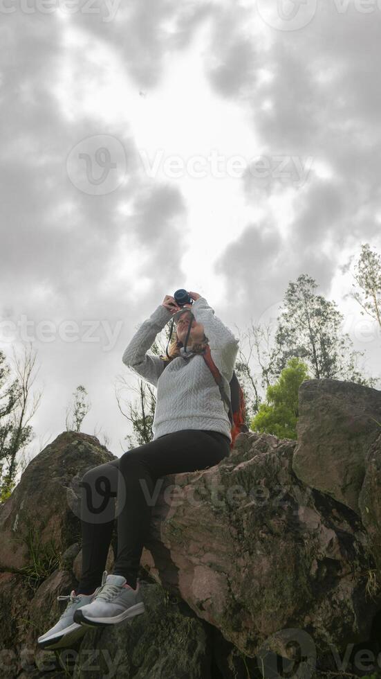 femme séance sur des pierres à le Haut de une Montagne prise une photo avec sa petit caméra sur une nuageux journée