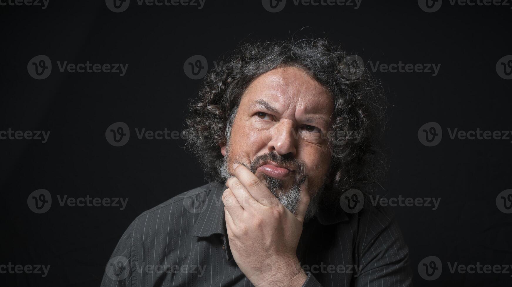 homme avec blanc barbe et noir frisé cheveux avec indécis expression, main sur menton, portant noir chemise contre noir Contexte photo