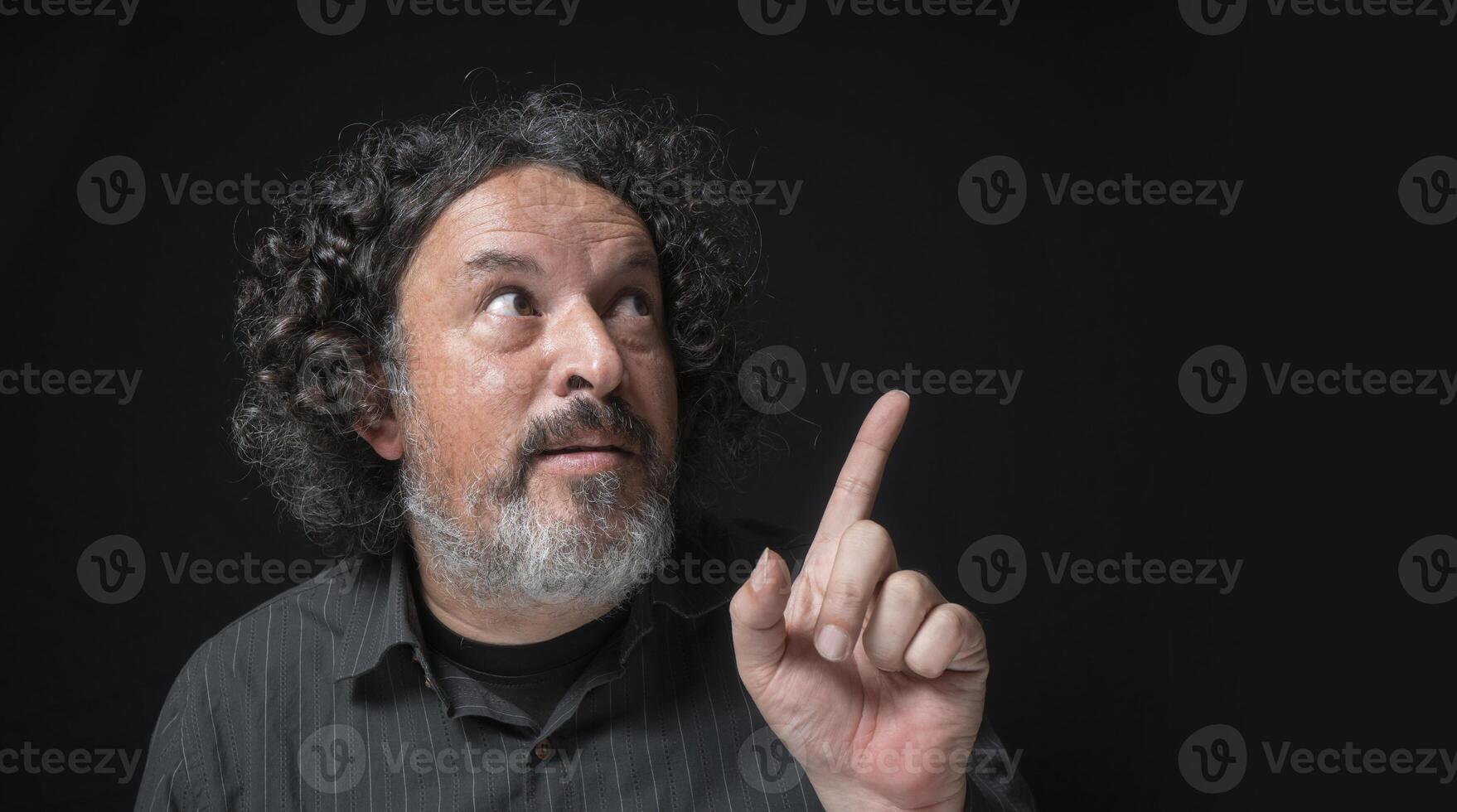 homme avec blanc barbe et noir frisé cheveux avec marrant expression, montrer du doigt en haut avec le sien main, portant noir chemise contre noir Contexte photo