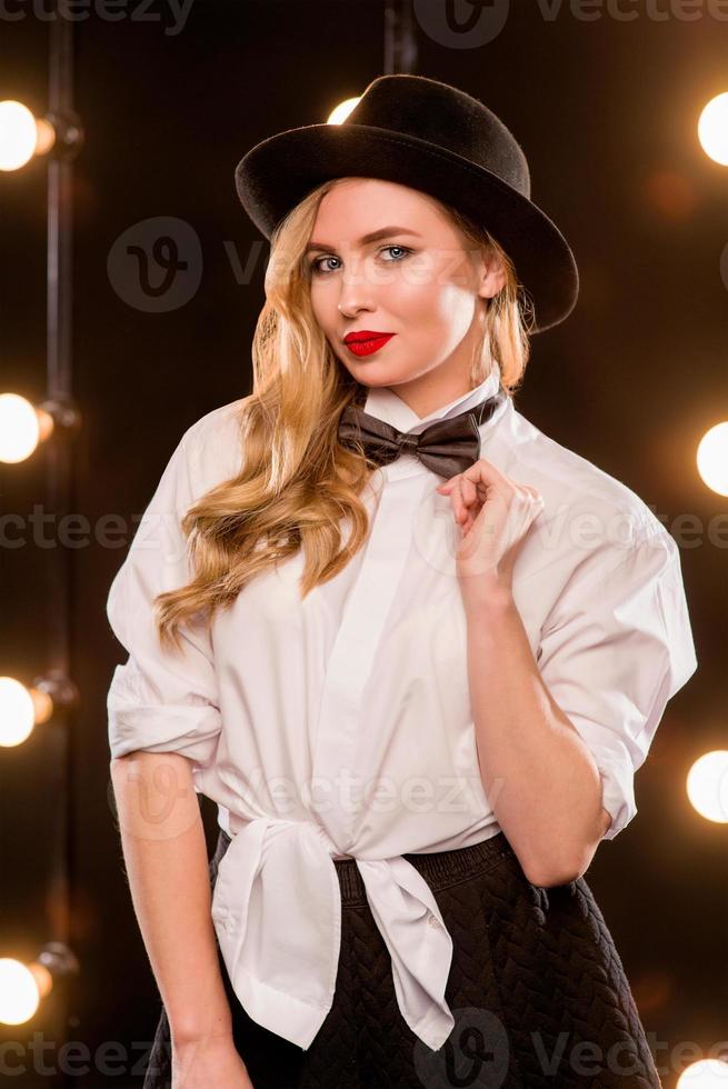 Jeune femme séduisante blonde en chemise blanche, papillon, chapeau noir avec microphone chantant sur scène photo