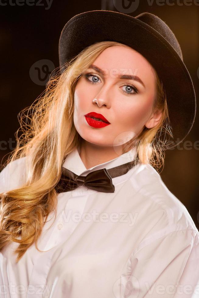 Jeune femme séduisante blonde en chemise blanche, papillon, chapeau noir avec microphone chantant sur scène photo