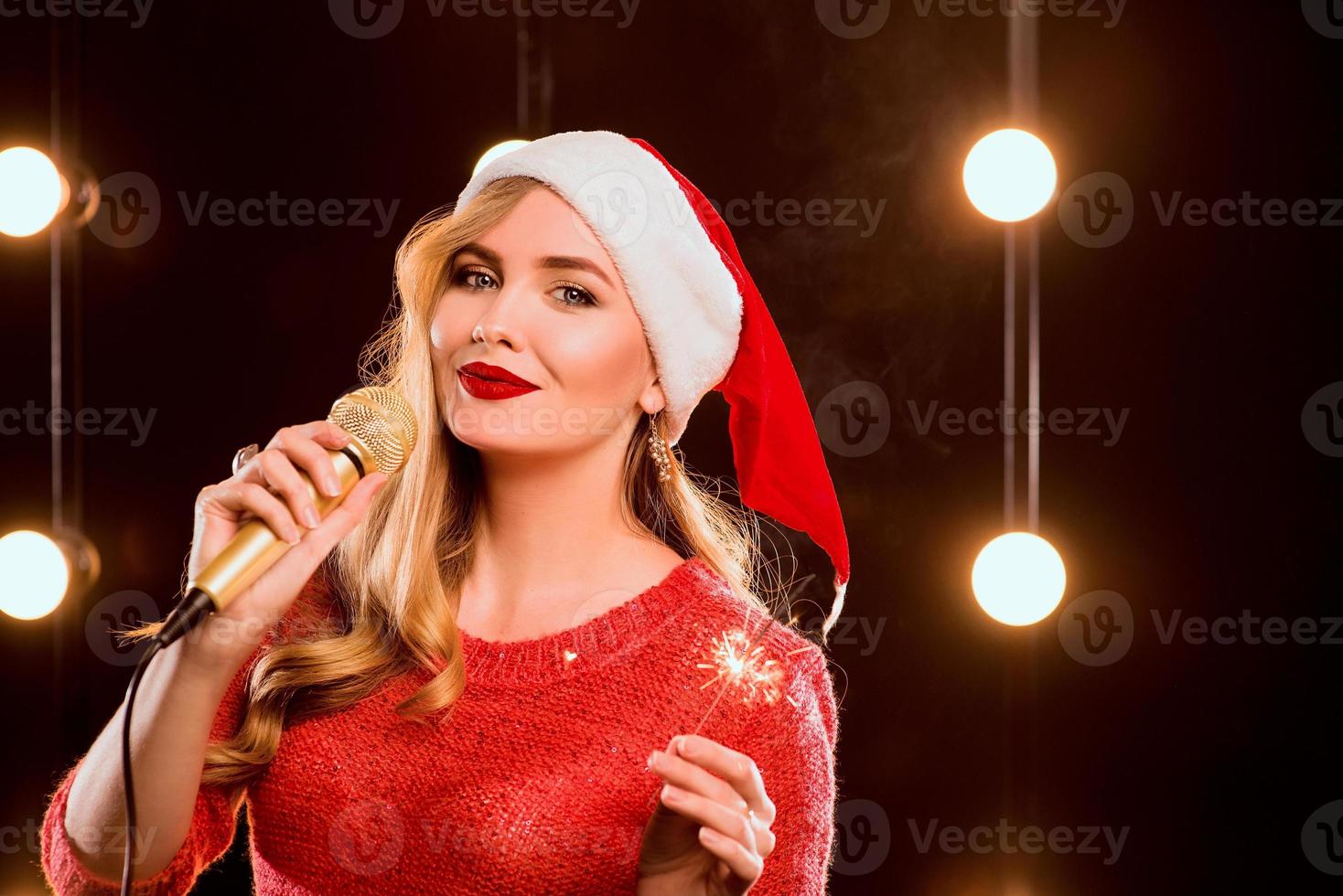 Jeune femme séduisante blonde en pull rouge et chapeau de nouvel an rouge avec microphone et cierge magique sur scène photo