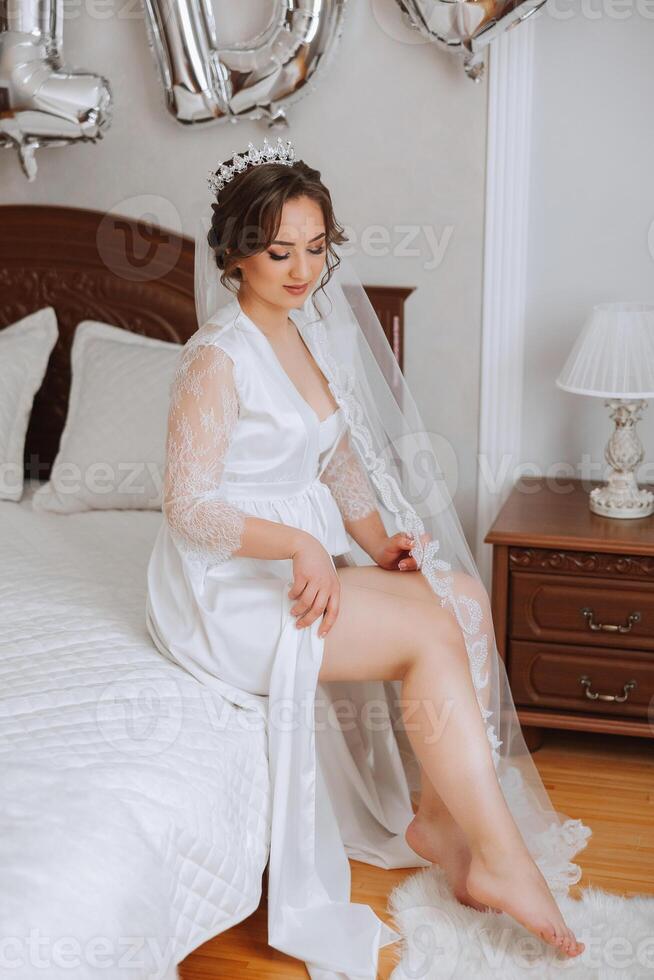 une magnifique brunette la mariée avec une tiare dans sa cheveux est avoir prêt pour le mariage dans une magnifique peignoir dans boudoir style. fermer mariage portrait, photo. photo