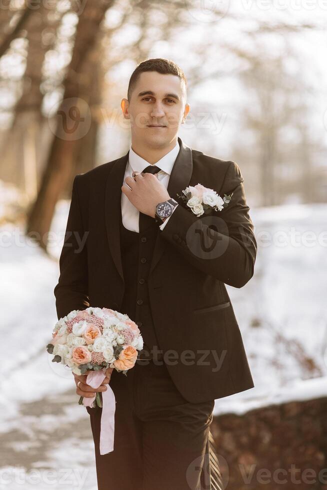 portrait de une Beau et viril jeune marié dans une noir costume avec une cravate. jeune marié dans la nature dans le hiver saison. photo