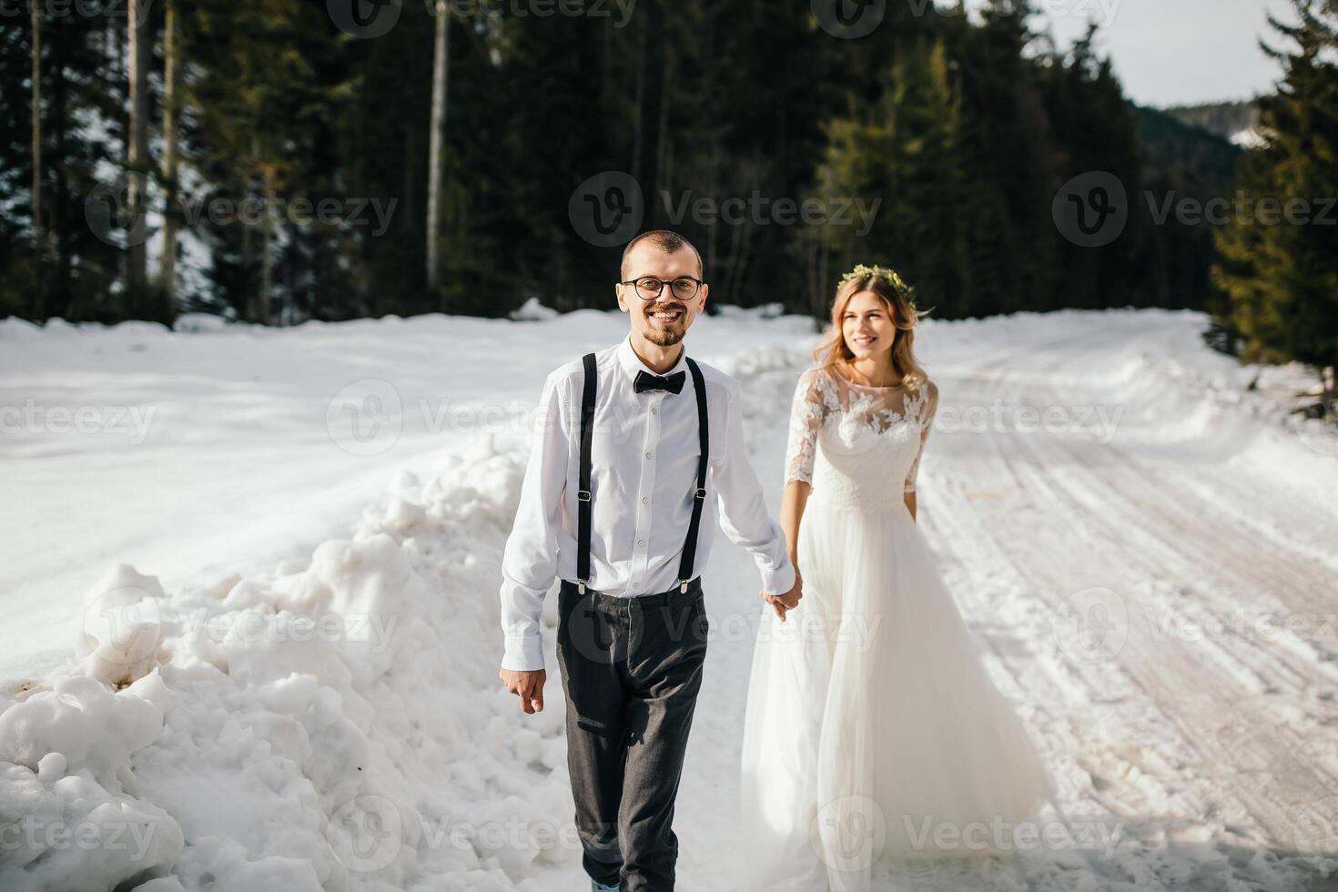 le la mariée et jeune marié sont fonctionnement le long de une neigeux route contre le Contexte de une pin forêt et magnifique contraste lumière du soleil. côté voir. hiver mariage. endroit pour logo. photo