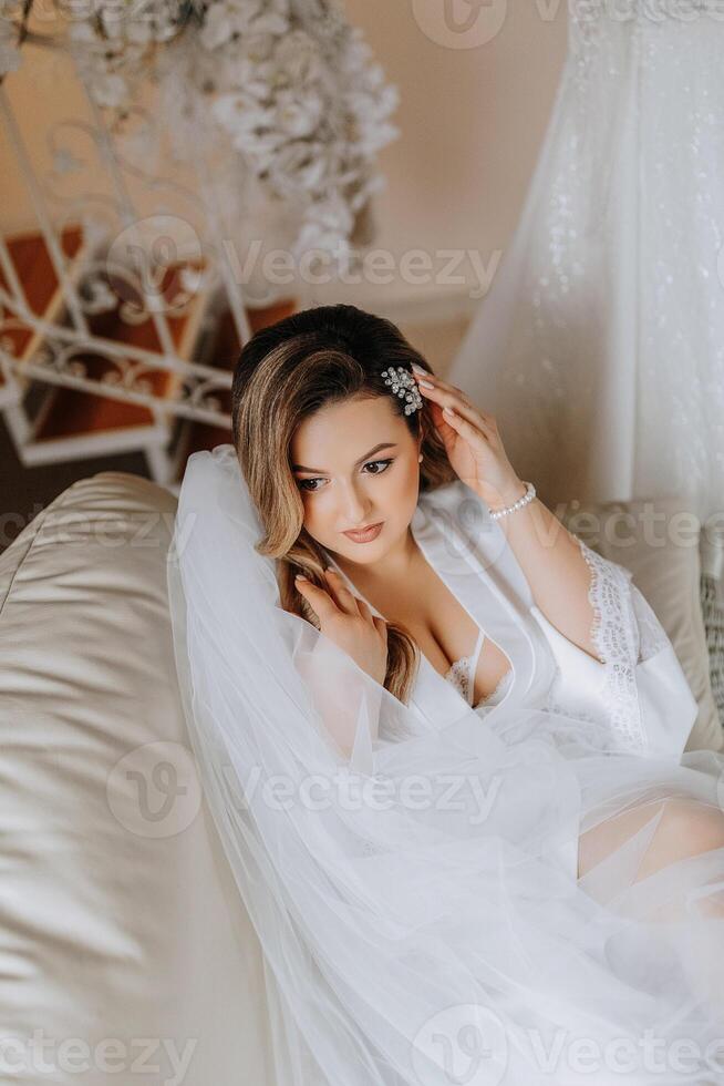 Matin de le la mariée avant le mariage. une magnifique Jeune femme avec une voile et une magnifique coiffure dans une blanc peignoir dans une pièce avec une magnifique intérieur. Naturel beauté et professionnel se maquiller. photo