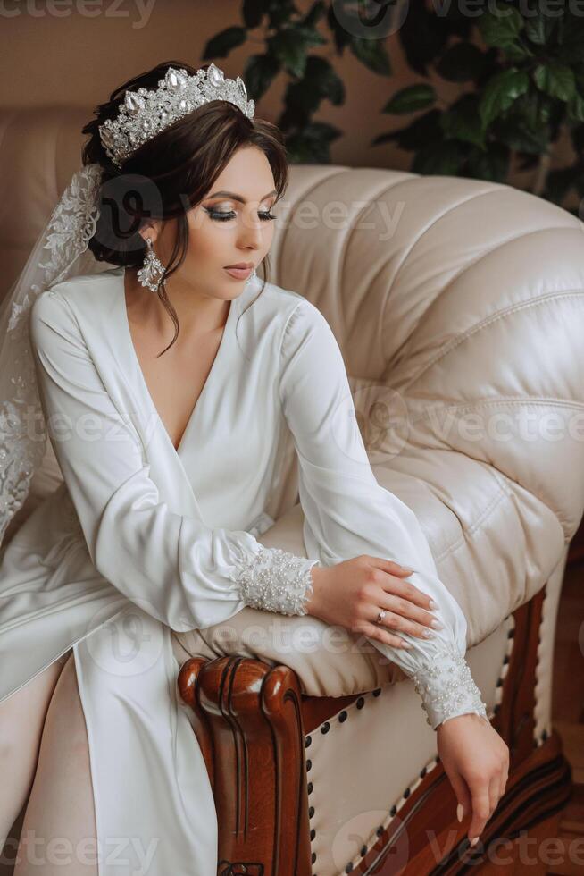 une magnifique brunette la mariée avec une tiare dans sa cheveux est avoir prêt pour le mariage dans une magnifique peignoir dans boudoir style. fermer mariage portrait, photo. photo