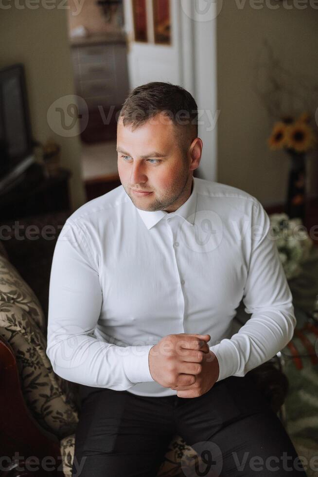 le jeune marié boutons le sien chemise par le la fenêtre. homme dans une chemise. Masculin portrait. séance photo à le la fenêtre. du marié collection. mariage journée. Beau homme jeune marié boutonnage le sien chemise dans une Hôtel chambre.