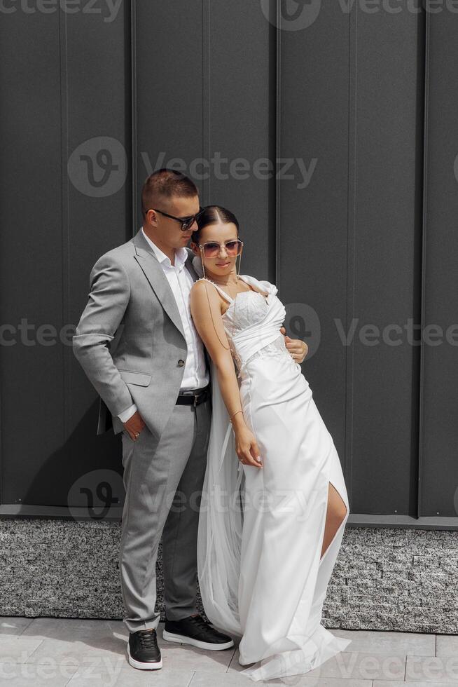 à la mode et mise en scène photo de le la mariée et jeune marié contre une gris mur. la mariée avec une longue voile. jeune marié et la mariée dans des lunettes de soleil.