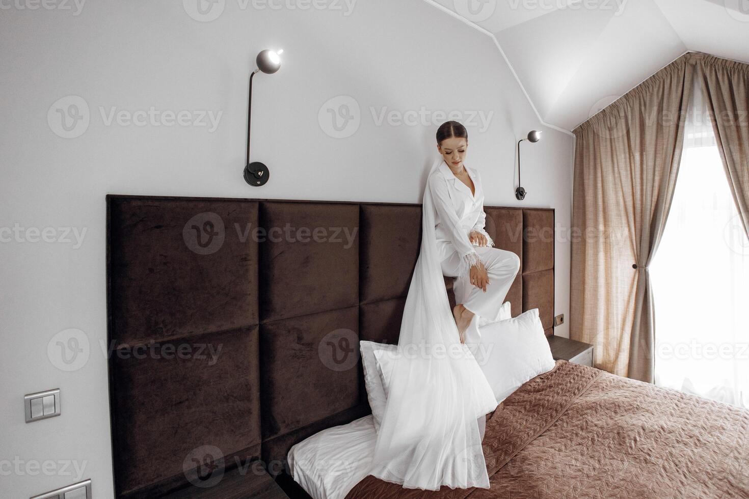 une magnifique et sur de soi la mariée permanent sur une lit dans blanc pyjamas avec une longue voile dans une Hôtel pièce pose pour une photographe. mariage concept. Matin de le la mariée. préparation pour le mariage. photo