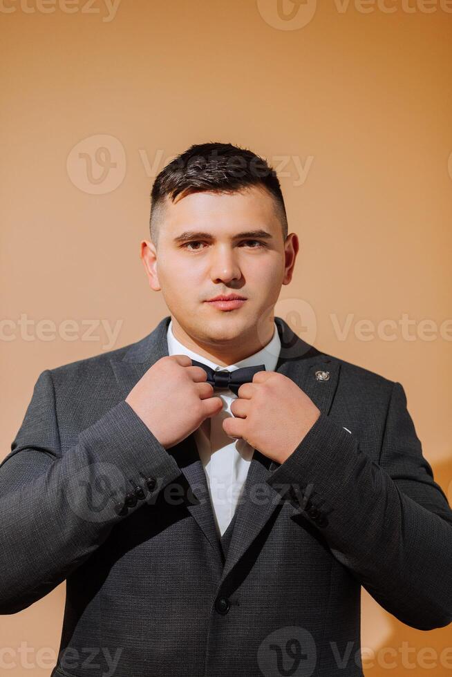 une Jeune souriant homme dans une blanc chemise porte une arc attacher et une noir gilet. une jeune marié avec une court coiffure sur un Orange Contexte met sur le sien noir costume et prépare pour le mariage photo