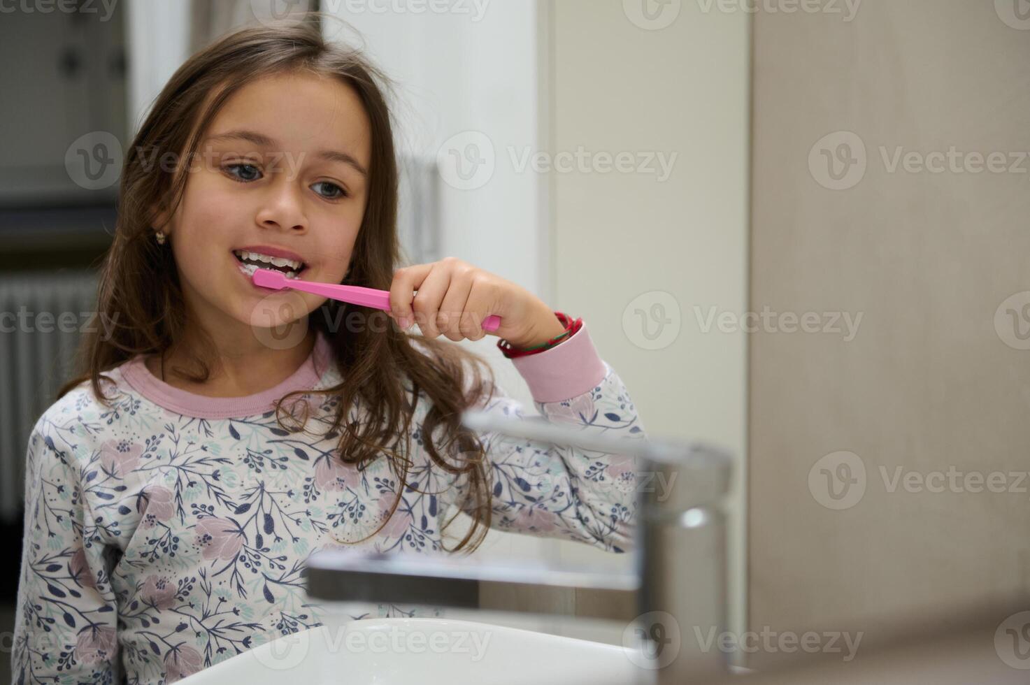 fermer authentique portrait de une caucasien mignonne enfant fille brossage dents, admiratif se dans le salle de bains miroir. photo