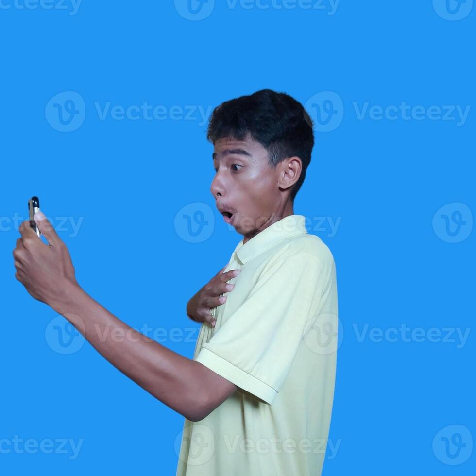 Jeune asiatique homme surpris à la recherche à intelligent téléphone isolé bleu Contexte. portant une Jaune T-shirt photo