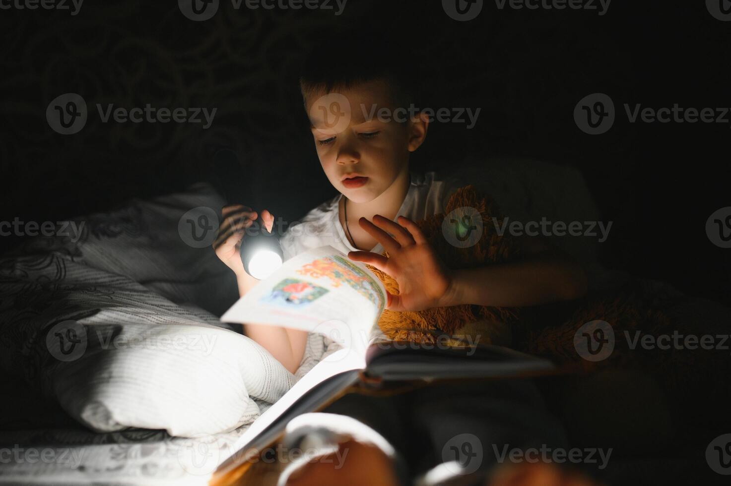 garçon avec lampe de poche en train de lire livre en dessous de couverture à maison. photo