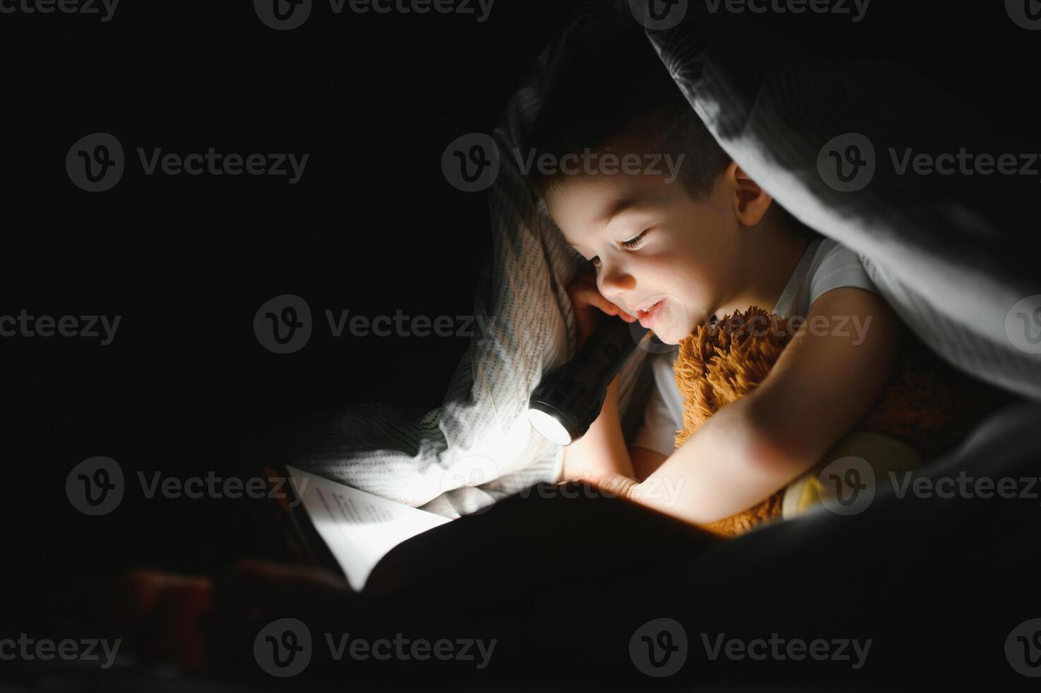 une garçon de 5-6 ans vieux est en train de lire une livre dans le soir dans le foncé en dessous de une couverture avec une jouet ours. photo