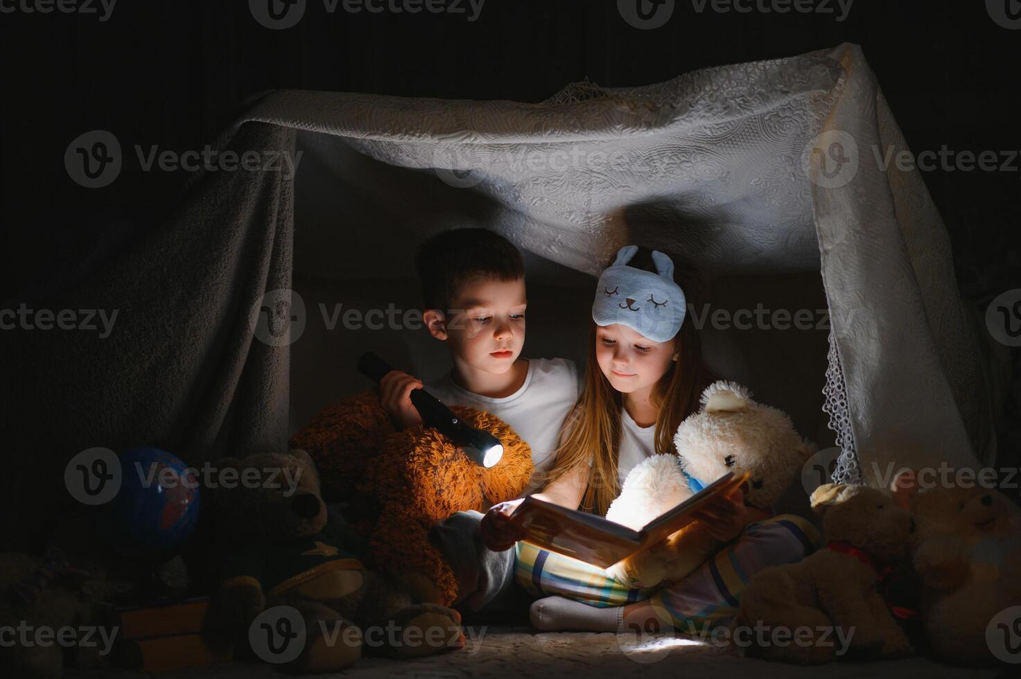 Fratrie asseoir dans une cabane de chaises et couvertures. frère et sœur en train de lire livre avec lampe de poche à maison. photo