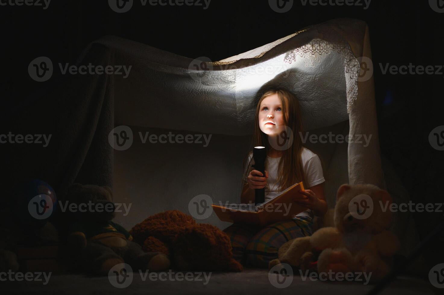 peu fille sont en train de lire une livre avec lampes de poche dans tente. content fille en jouant à maison. marrant charmant enfant ayant amusement dans pièce photo