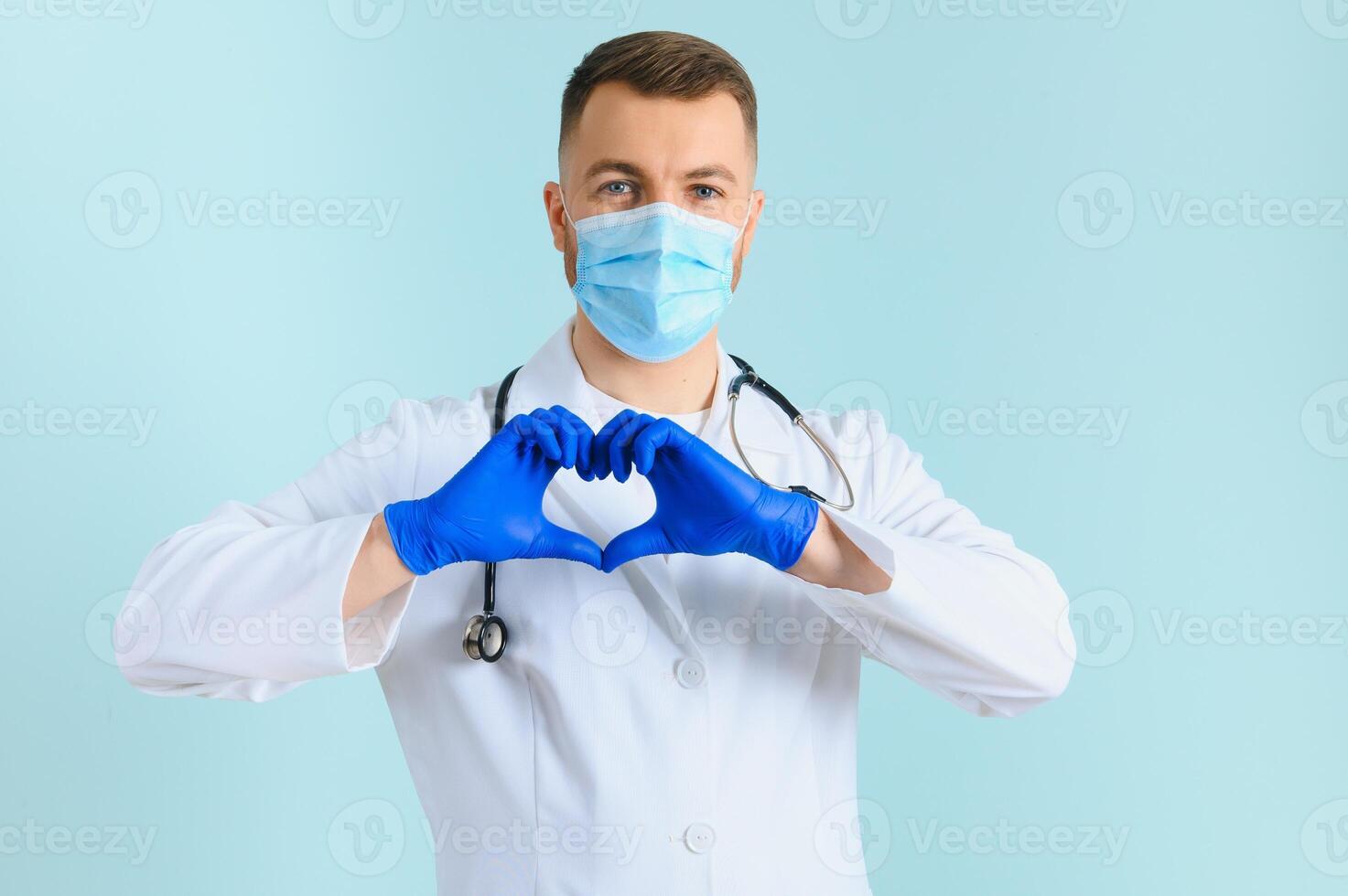 médecin mains dans gants dans le cœur forme contre le Contexte de le sien corps et médical robe. fermer. Masculin main dans bleu médical gants spectacle cœur signe photo